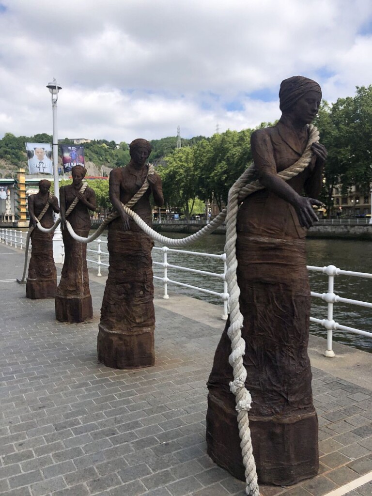 Zdjęcie pomnika przedstawiającego cztery postacie kobiece połączone ze sobą liną okrętową. w tle rzeka i domy przy kamiennym nabrzeżu