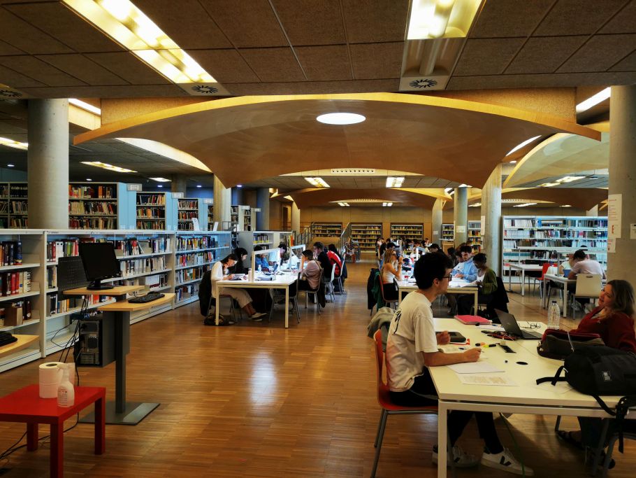 Zdjęcie czytelni bibliotecznej. Ludzie siedzący przy stolikach i rzędy półek z książkami.