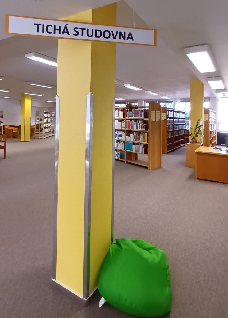 wnętrze z regałami pełnymi książek, na środku żółty słup z napisem: ticha studovna