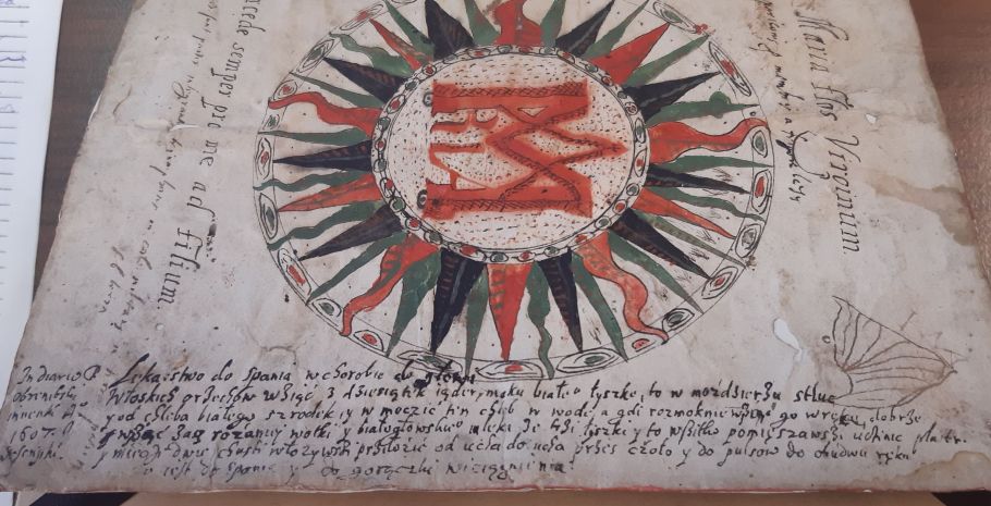 ilustracja z książki: okrąg otoczony zielonymi, czerwonymi i czarnymi promieniami, z wpisanymi literami MARIA
