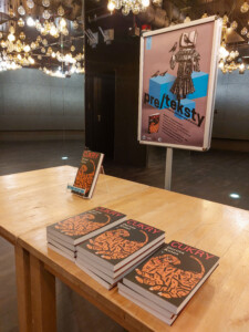 Widok na stół z egzemplarzami książki "Cukry", w tle plakat Pre/teksty na stojaku.