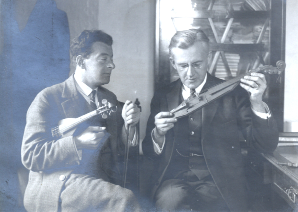 Zdjecie przedstawiające dwóch siedzących mężczyzn w garniturach. Przyglądających się instrumentom muzycznym - smyczkowym