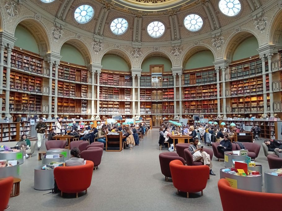 Ogromna czytelnia biblioteczna, na środku owalnego pomieszcenia fotele i niskie stoliki, wokół kilka pięter galerii z regałami zastawionymi książkami.