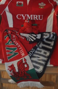 Pluszowa maskota w kształcie smoka, szalik z meczu Walia-Szkocja na tle czerwonej koszuli z napisem Cymru.