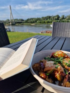 Zdjęcie otwartej książki i talerza zjedzeniem, w tle trawa, most i rzeka