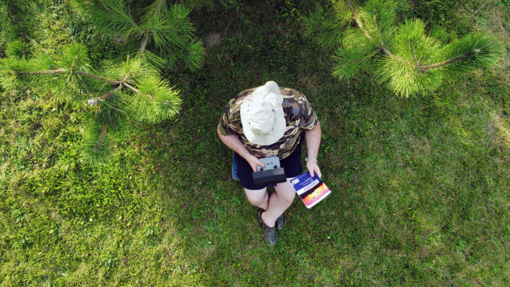 Widok z góry na człowieka siedzacego obok sosny i czytającego książkę i trzymającego radio rc i
