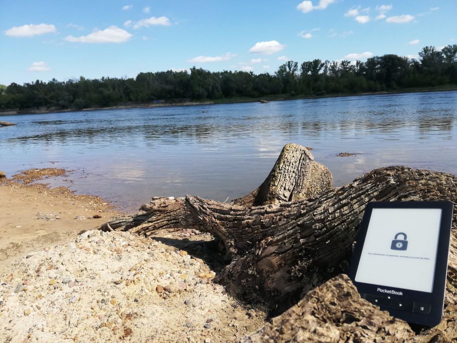 Zdjęcie rzeki, na pierwszym planie wyrzucone na brzeg drzewo i ułożony na nim e-czytnik.